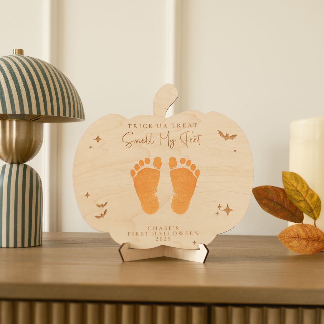 Wooden pumpkin-shaped plaque with footprints and text on a shelf with decorative items.